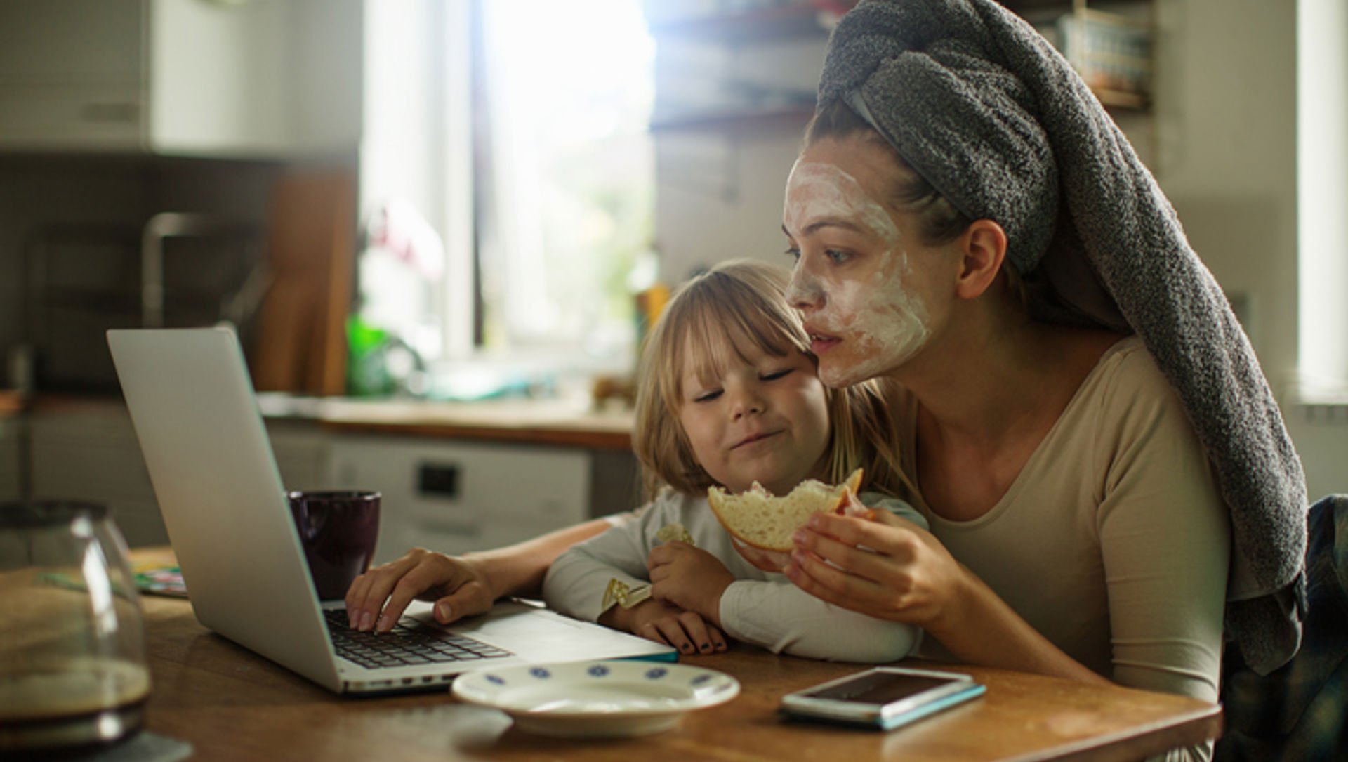 Mum and child having breakfast at the table while mum works on her laptop, face mask on and hair in a towel, with the child in her lap