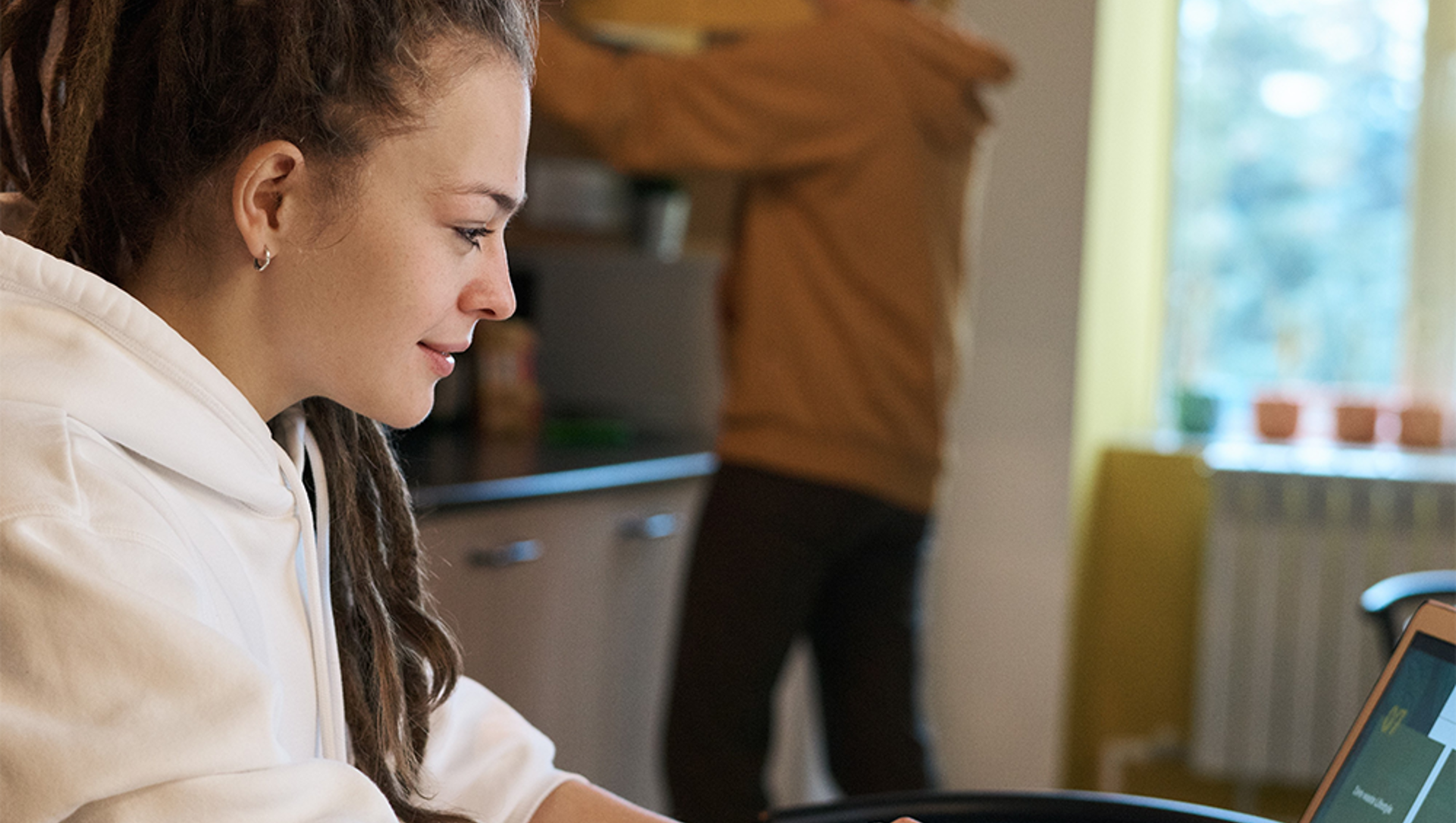 Woman sitting at the kitchen table on her laptop, man blurred in the background fetching something from the cupboard